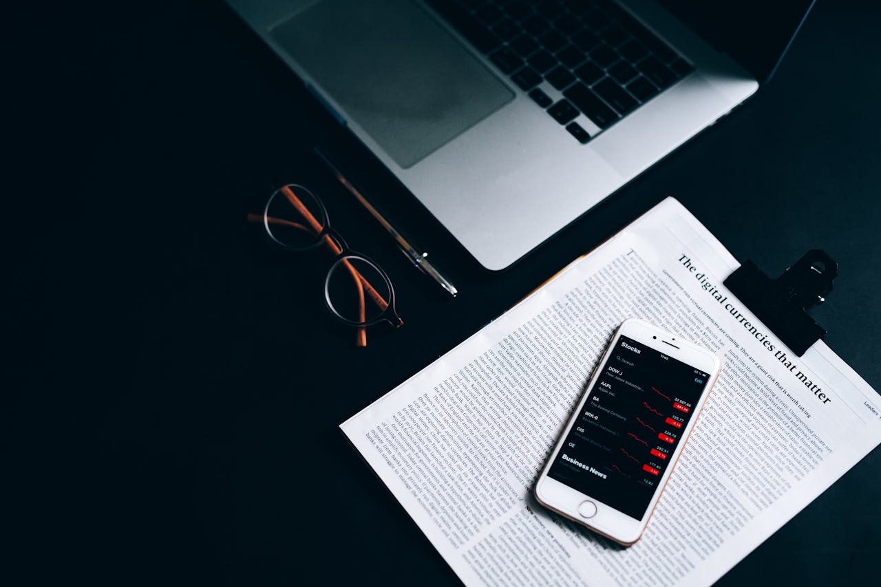 Flat lay of a digital workspace featuring a smartphone, glasses, and a laptop on a dark desk.